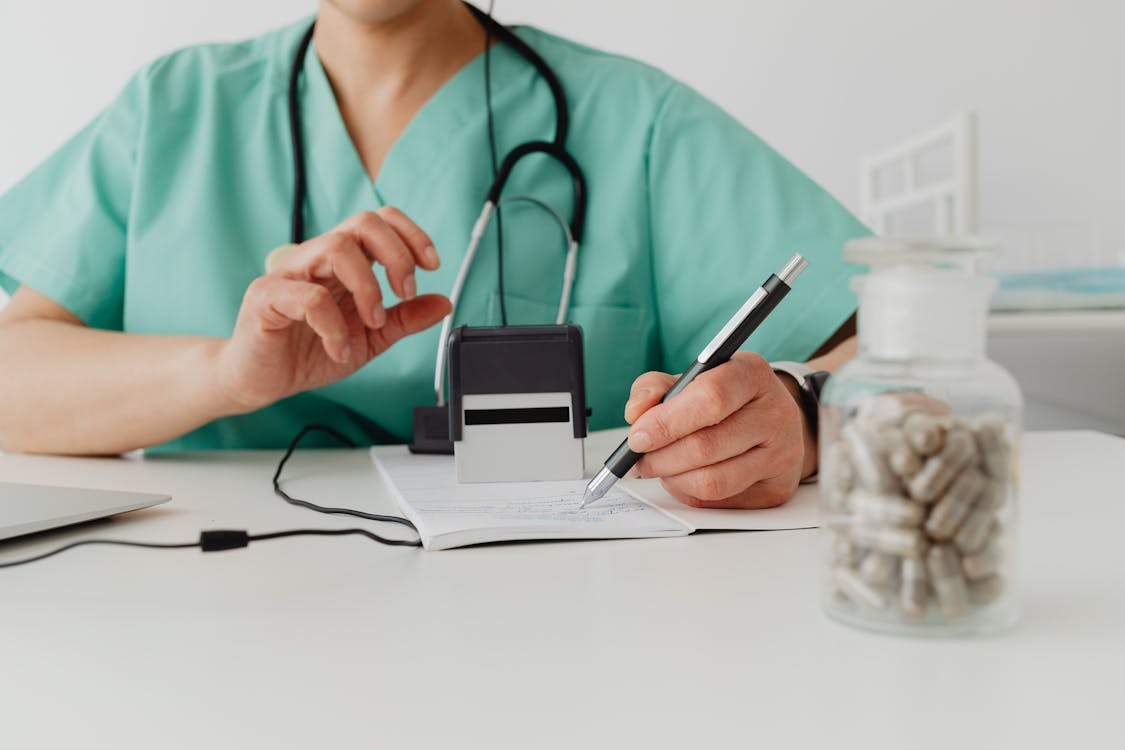 The image shows a healthcare professional in a green scrub top, writing notes on paper while using a stamp and having a jar of capsules visible on the table.