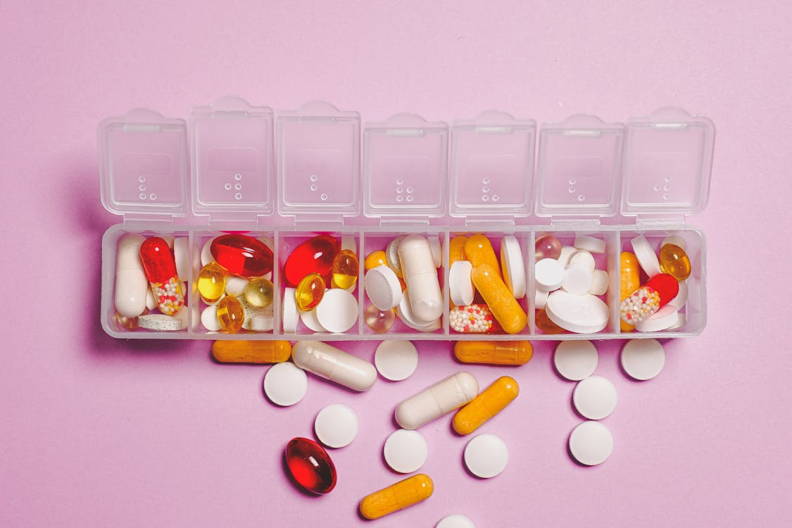 The image shows a weekly pill organizer filled with various colorful capsules, tablets, and soft-gel pills. Some pills are also scattered around the container on a pink surface.