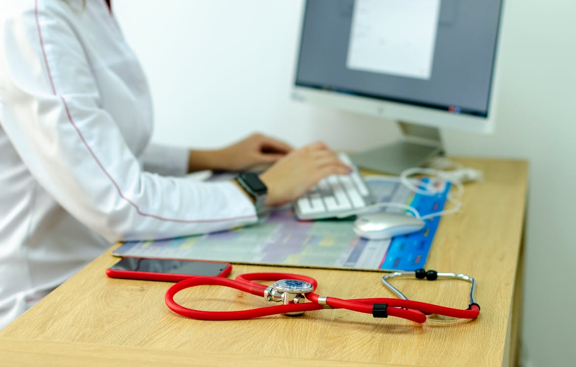 The image shows a healthcare professional typing on a computer, with a red stethoscope and a smartphone on the desk.