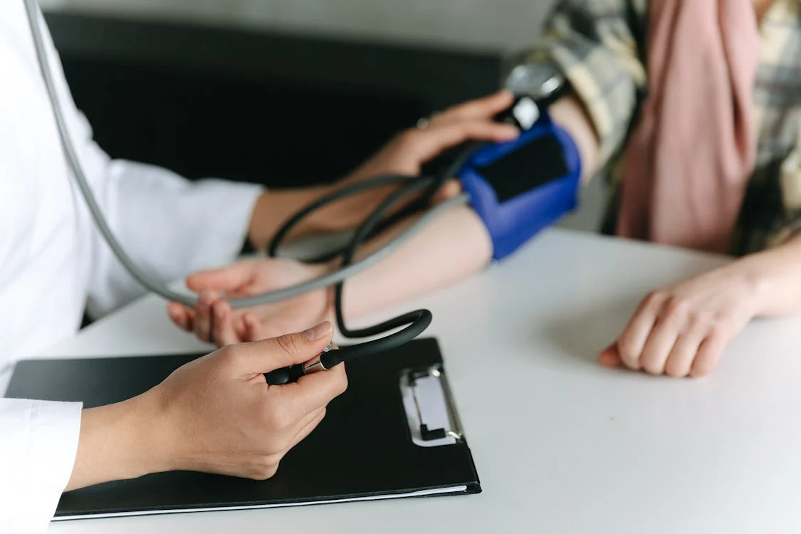 The image shows a medical professional measuring a patient's blood pressure using a stethoscope and a blood pressure cuff.