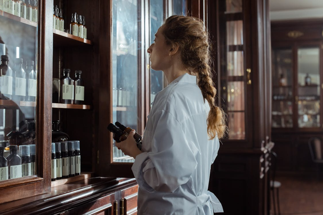 The image shows a woman in a lab coat, standing in front of a wooden shelf filled with bottles. She is holding a couple of bottles while inspecting the shelf.