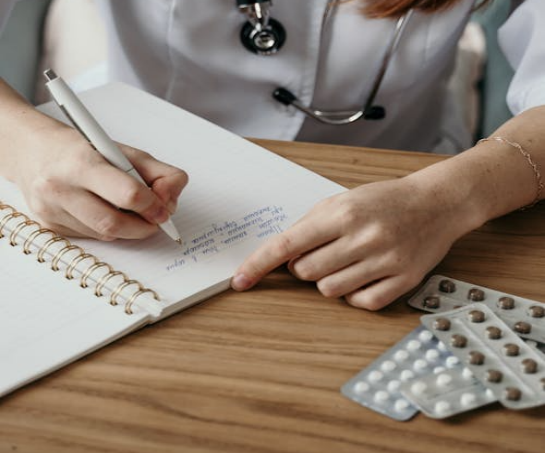 The image shows a medical professional writing notes in a notebook on a wooden table beside several packs of medication tablets.