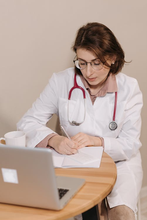 Female doctor writing notes beside a laptop during a consultation