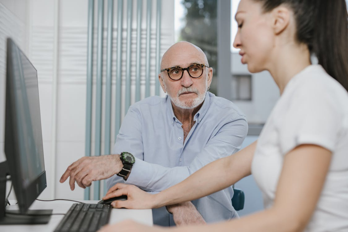 Older man seated at a desk reviewing information on a computer with another person.