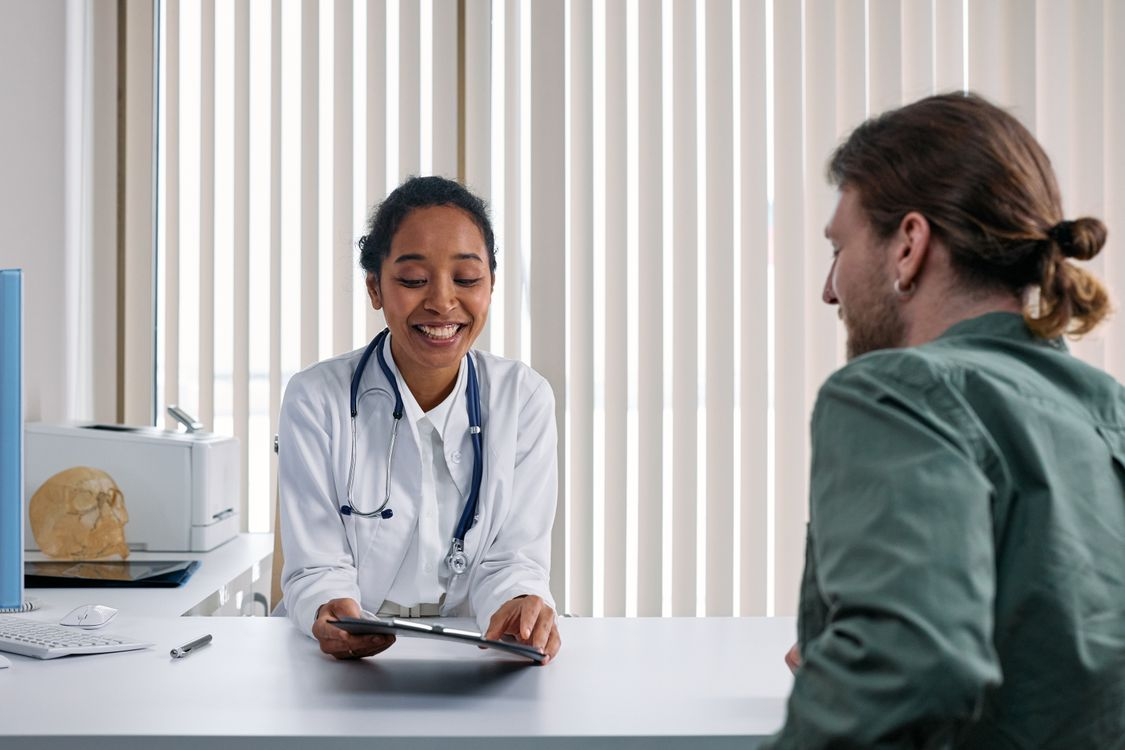 A smiling female doctor in a white coat consulting with a male patient at her desk.