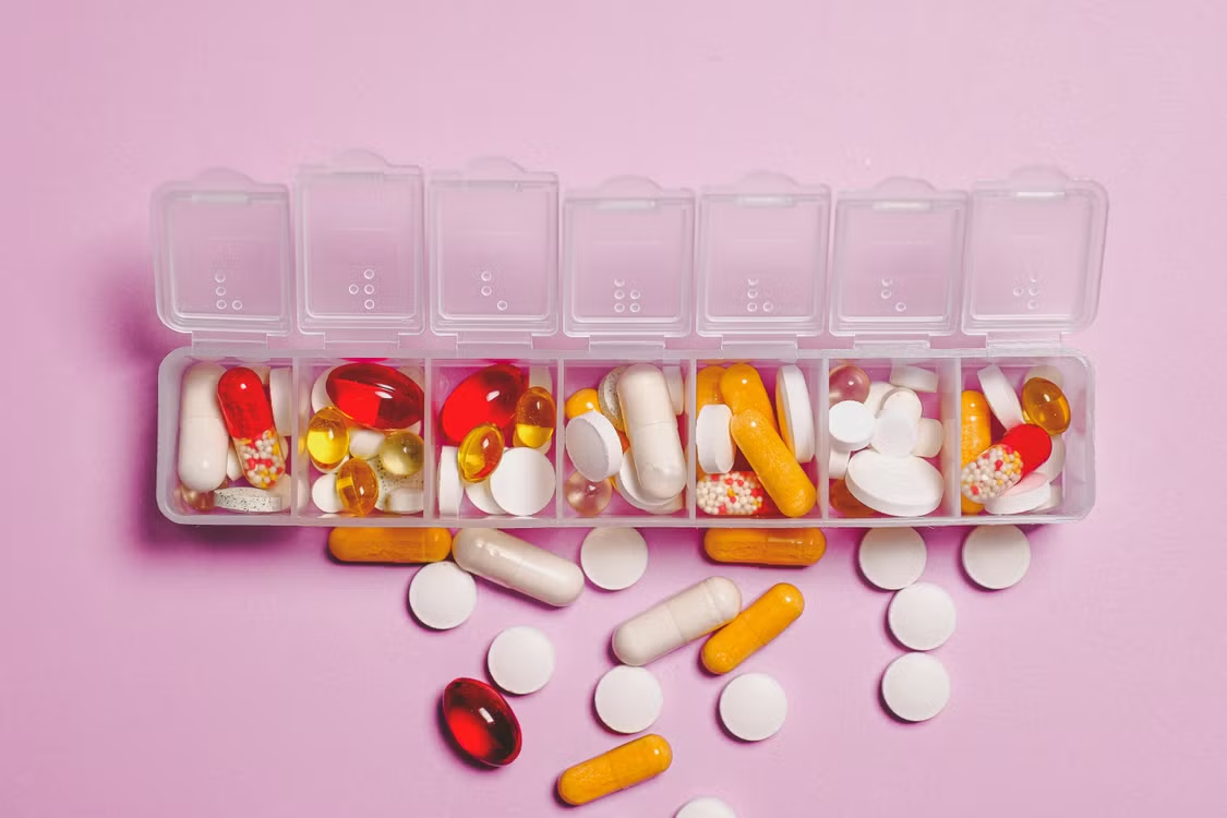 A weekly pill organizer filled with assorted colorful medications on a pink background. Caption: Patient assistance programs help uninsured and low-income patients afford the
