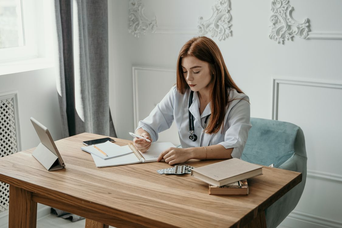 A female doctor in a white coat writing notes at a desk with prescription medications and a tablet nearby.