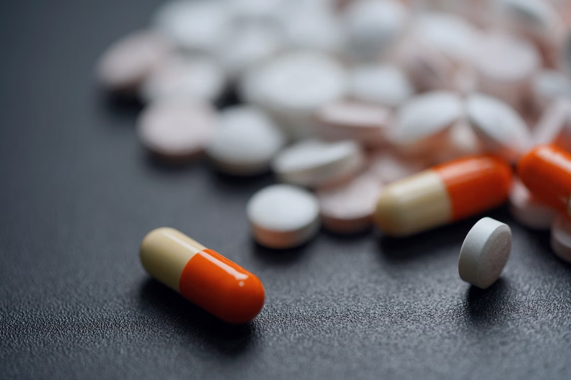 Orange-and-beige capsules and assorted white and pink tablets scattered across a dark surface.