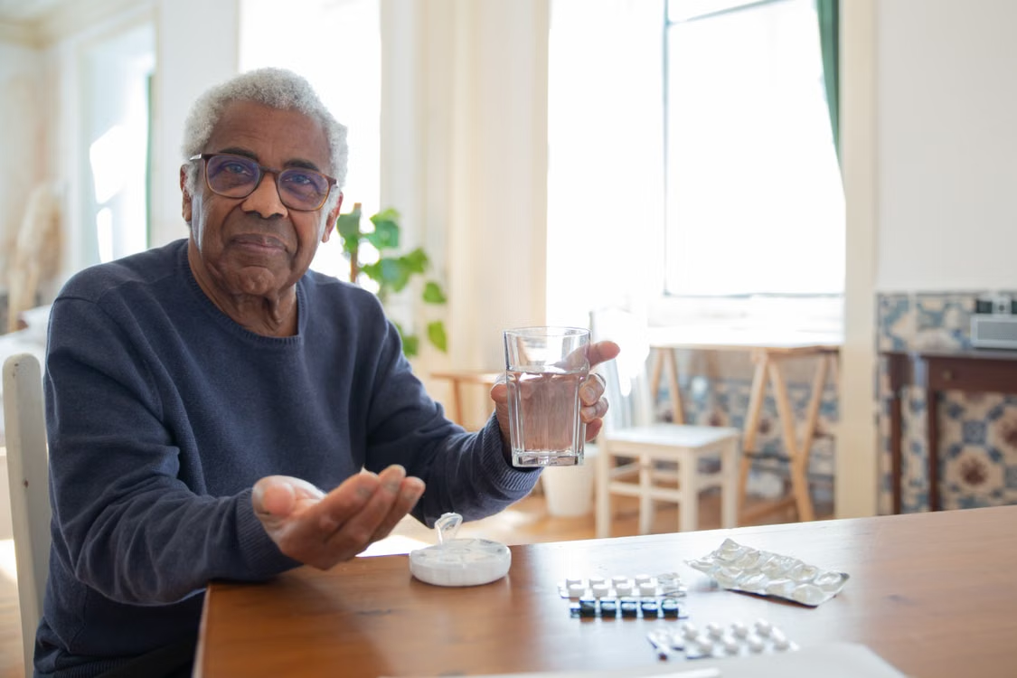 Older man holding a glass of water and medication while sitting at a table with prescription tablets.
