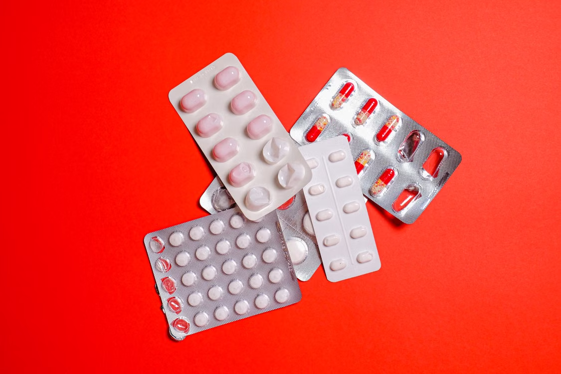 Several blister packs containing pink, white, and red tablets and capsules arranged on a bright red surface.