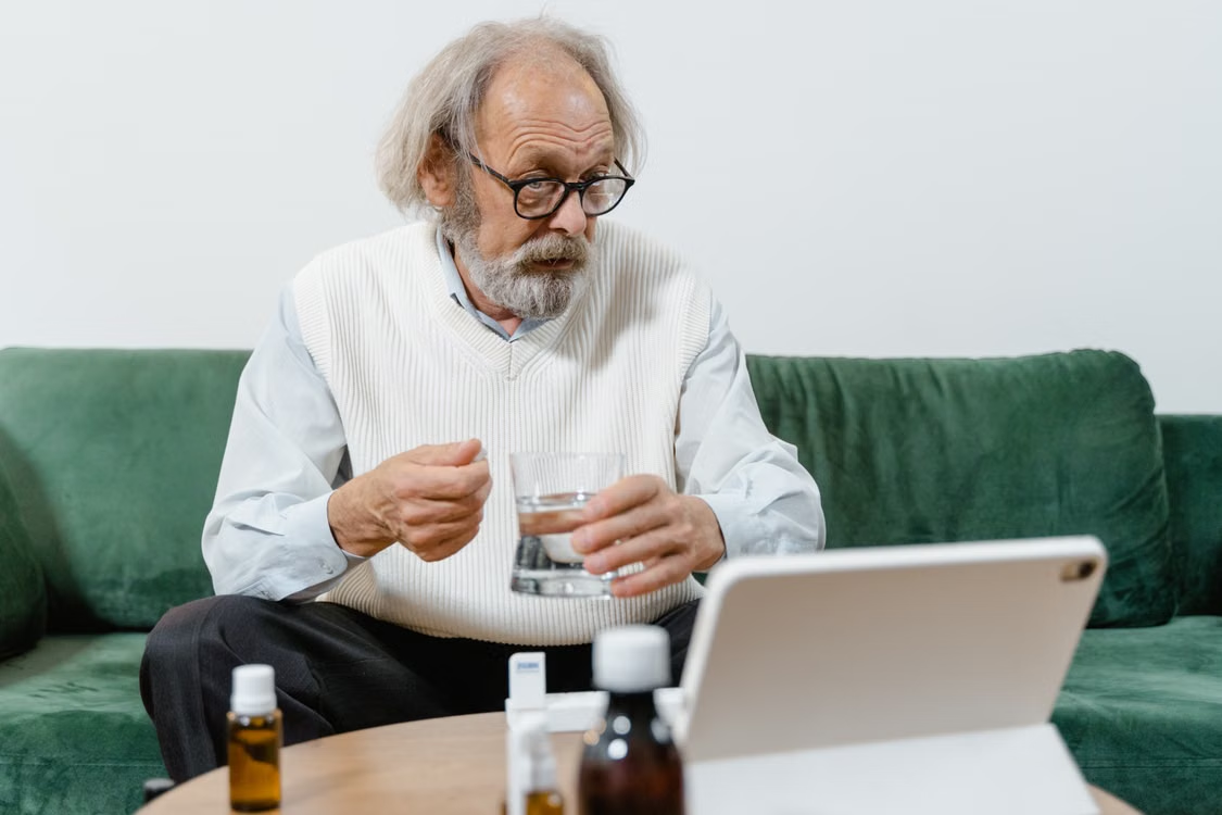 Older man holding a glass of water and medication while using a tablet at home.