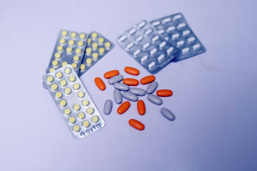 Yellow tablets in blister packs and loose orange and lavender capsules arranged on a light purple surface.