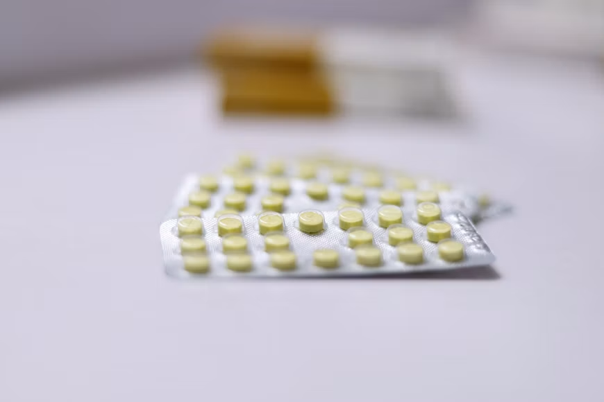 Yellow tablets arranged in a silver blister pack on a light-colored surface, with a blurred medication package in the background.