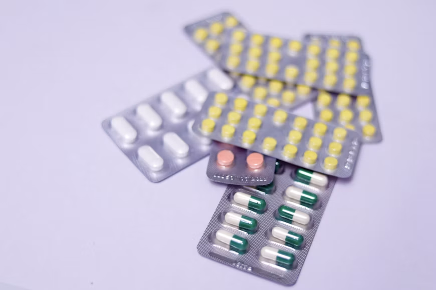 Blister packs containing yellow, white, pink, and green-and-white tablets arranged on a light purple surface.