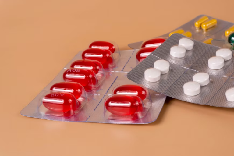 Red capsules, white tablets, and yellow capsules displayed in overlapping blister packs on a peach-colored surface.
