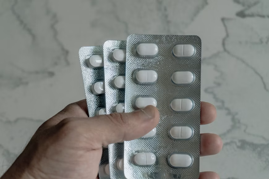 Hand holding three silver blister packs containing white tablets against a light marble surface.