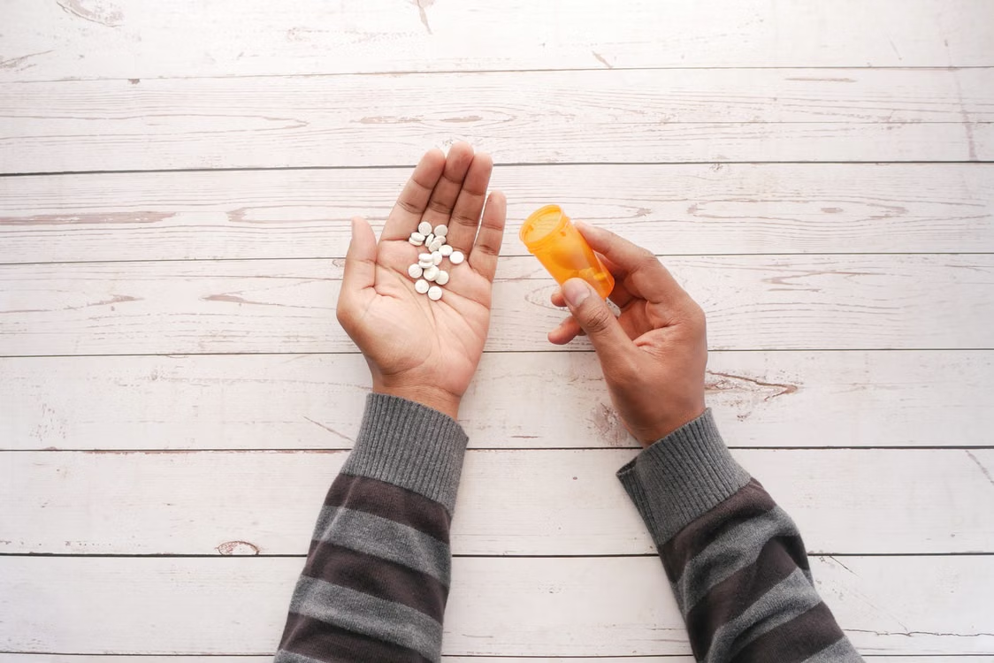 Person holding white tablets in an open palm while tipping an orange prescription bottle over a light wooden table.