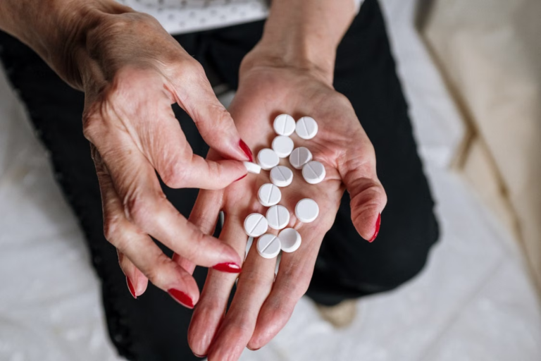 Older adult holding several white prescription tablets in their hands while seated at home.